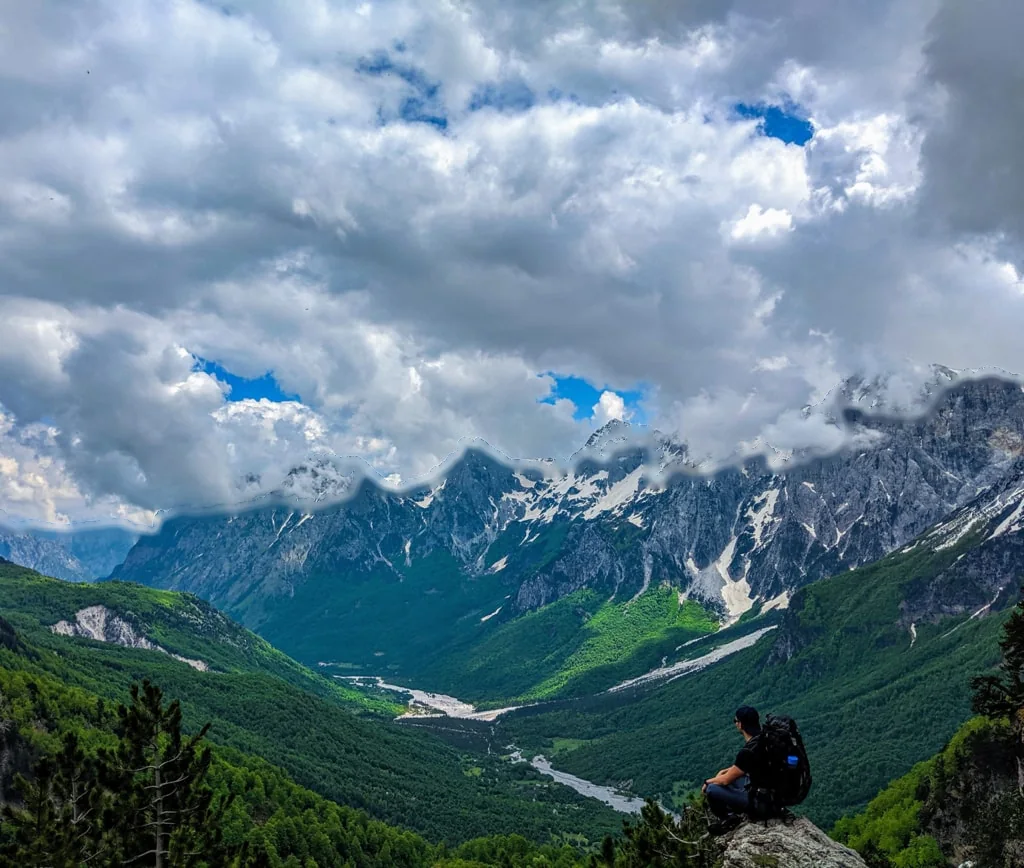 Ein Wanderer sitzt über dem grünen Valbonë-Tal, dahinter Kalkgipfel und Wolken