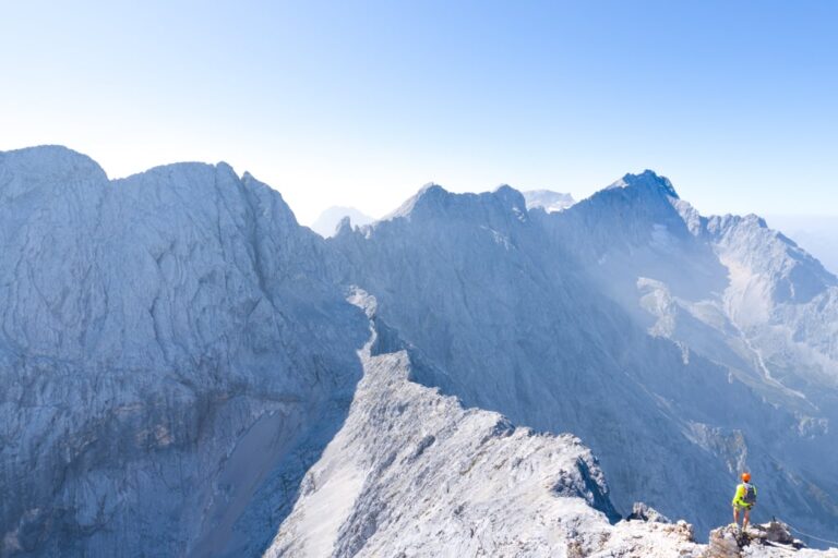 A hiker stands on a snowy ridge above steep Alpine peaks near the Alpspitze in clear morning light