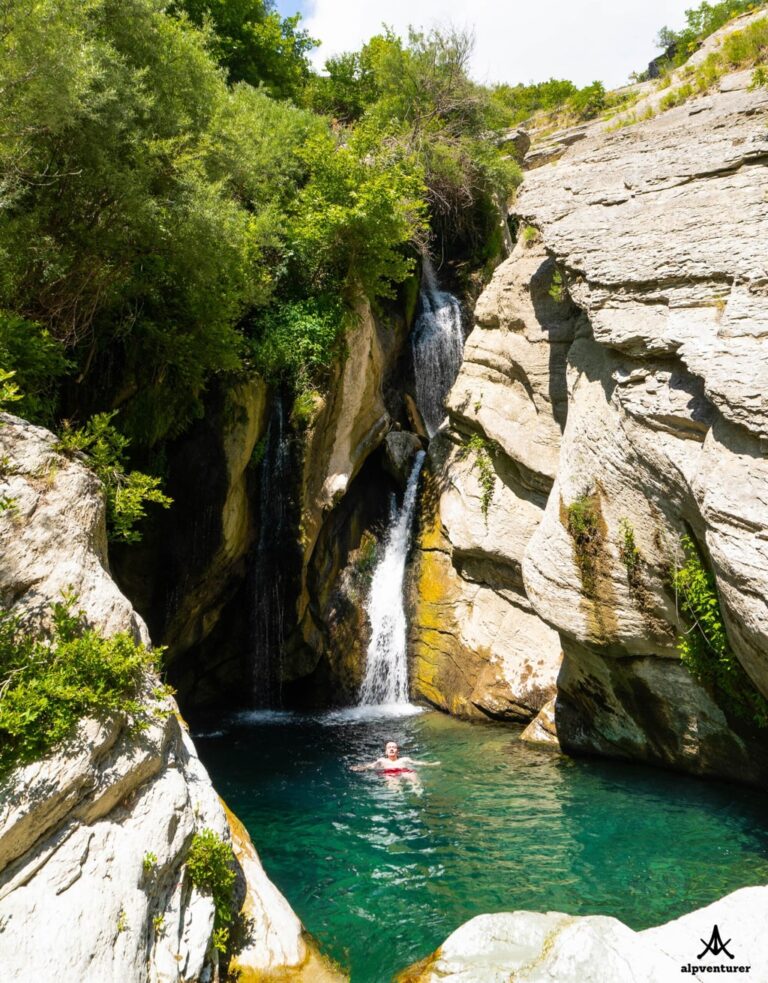 Bogova Waterfall drops between pale rocky cliffs into a turquoise pool where a swimmer floats