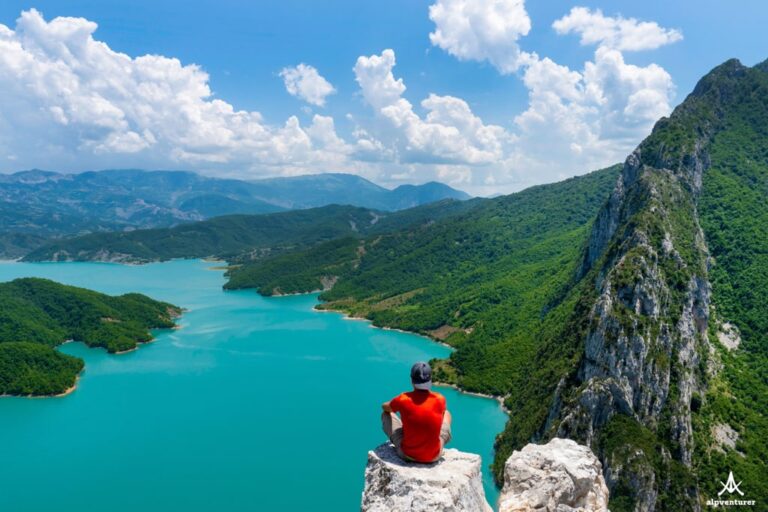 A hiker in a red shirt overlooks turquoise Lake Bovilla and green mountains under a cloudy blue sky