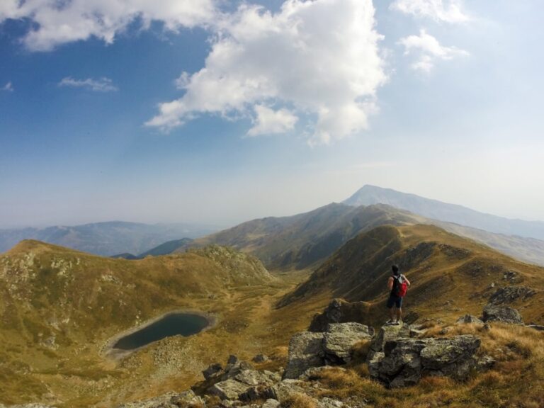 A hiker on a rocky ridge above Livadh lake, with the Luboten peaks under a partly cloudy sky
