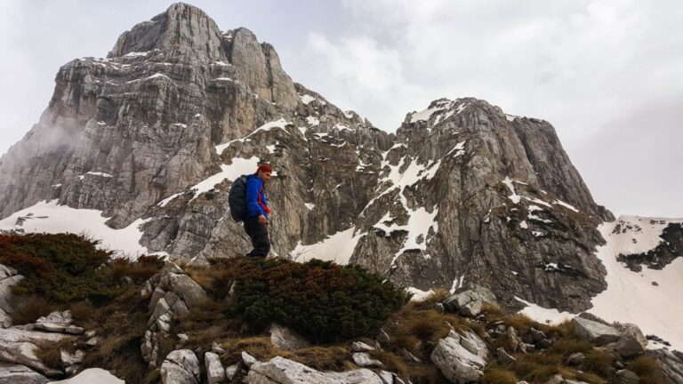 A hiker stands before snow streaking the rocky peaks of Cet Harusha in Albania