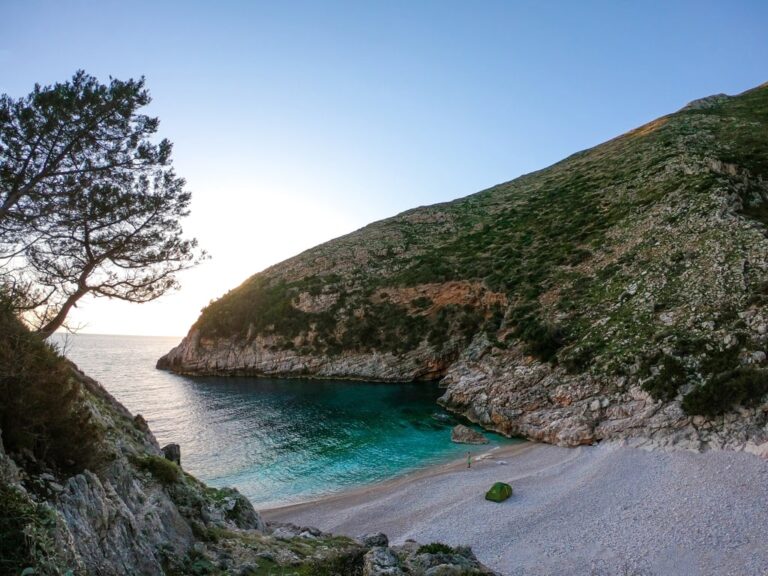 Dafina Bay’s turquoise water and pebbled shore beneath rocky hills in the soft light of late afternoon