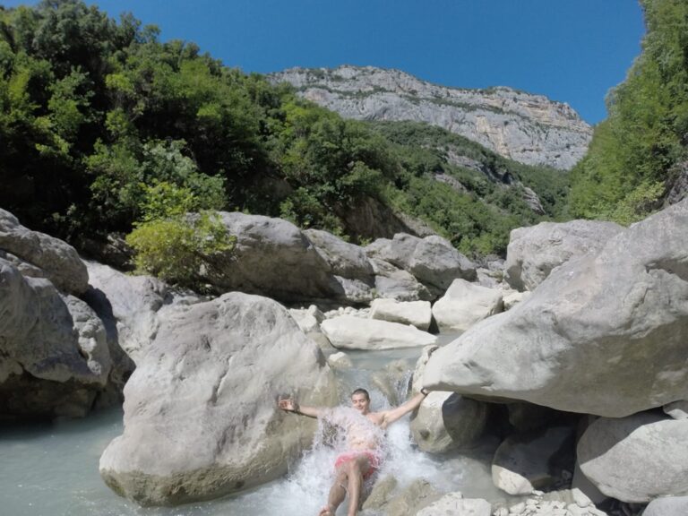 A hiker splashes through the Erzen River among pale boulders beneath a rocky, forested canyon ridge