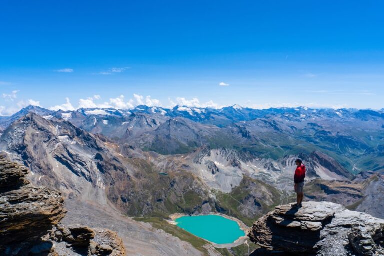 A hiker overlooks a turquoise alpine lake and rugged French Alps beneath a clear blue sky