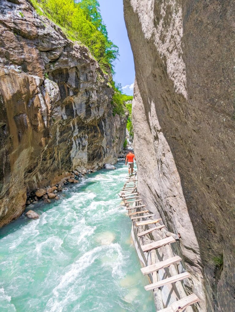 A hiker crosses a narrow wooden walkway above the Gashi River between steep gorge walls