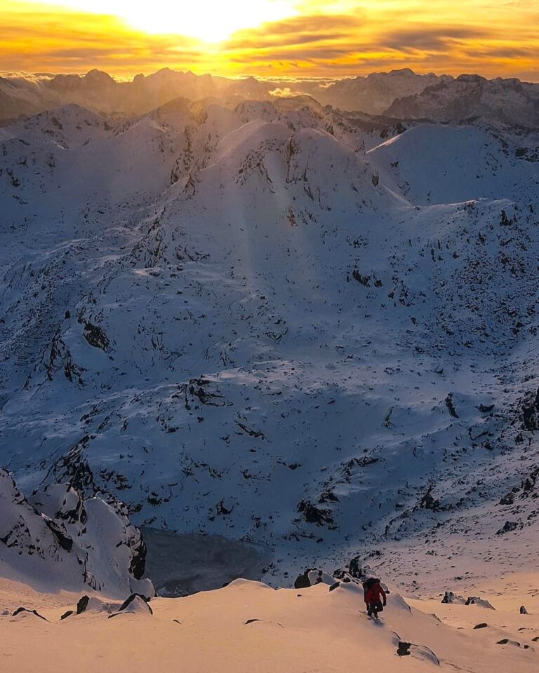 Snow-covered Accursed Mountains seen from Gjeravica at sunset, with a hiker descending a steep slope
