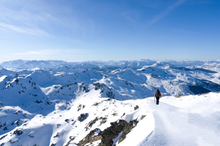 A hiker crosses a snowy ridge on Gjeravica Peak, with snow covered mountains beneath a clear blue sky