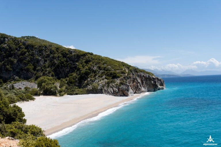 Sandy Gjipe Beach curves beneath a green rocky headland beside clear turquoise water in bright daylight