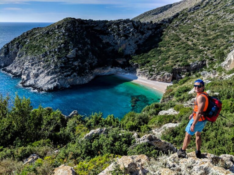 A hiker above Grama Bay, with turquoise water, a narrow sandy beach, and rocky green hillsides