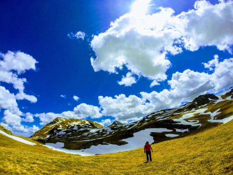 A hiker crosses grassy alpine slopes below snow patches on Great Rudoka under a cloud-filled blue sky