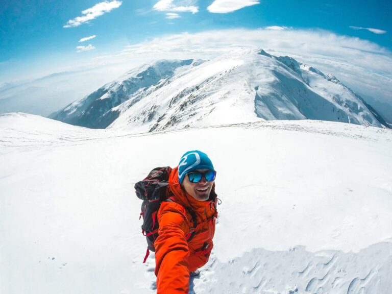 A hiker in orange gear crosses a snowy ridge in the Sharr Mountains beneath a blue sky