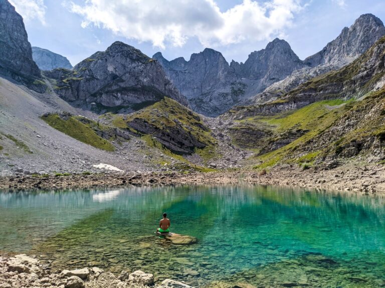 A hiker sits in a turquoise Jezerca lake beneath rocky Albanian peaks in bright daylight