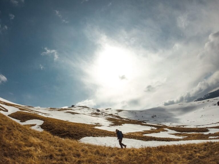 A hiker crosses snowy slopes on Bjeshka e Kabashit beneath a bright, clouded winter sky