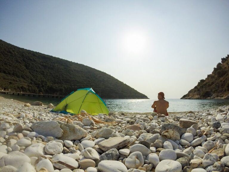 A tent and hiker on the rocky beach at Kakome Bay, with calm water and wooded hills in bright sunlight
