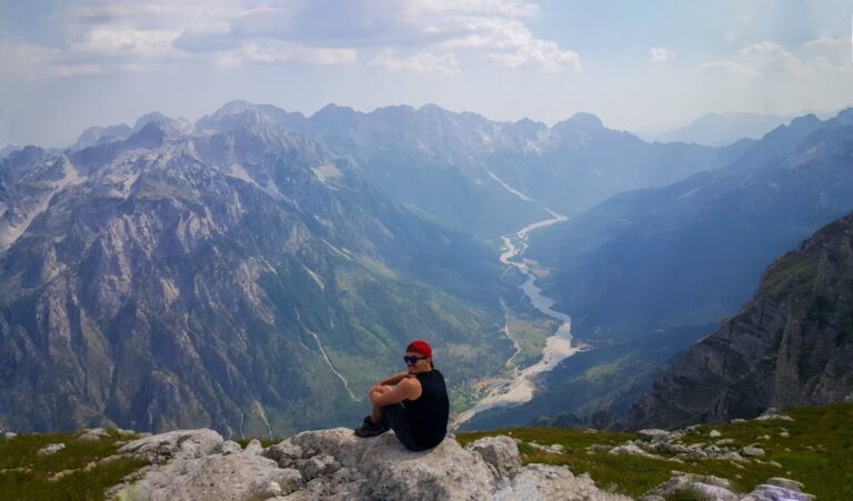 A hiker sits on a rocky summit overlooking the Valbona Valley and its rugged mountain ranges