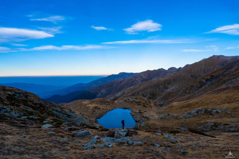 A hiker stands beside Konjusha Lake in a rocky mountain basin beneath a blue sky