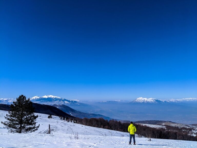 A hiker in a bright yellow jacket crosses a snowy ridge at Kopana Voda beneath distant snowcapped mountains