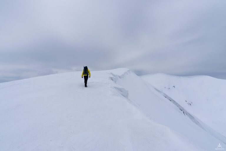 A hiker in a yellow jacket crosses a broad, snow covered ridge on Koritnik beneath a cloudy sky