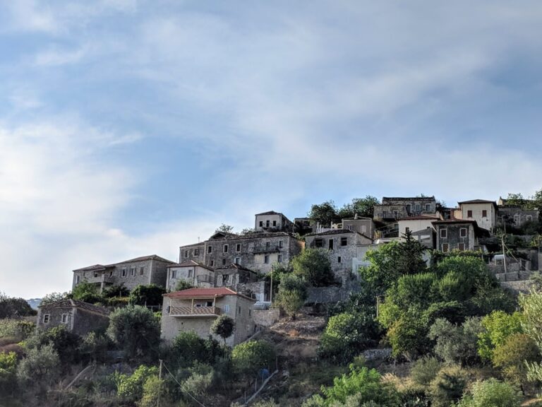 Stone houses of Kudhës village scattered across a green hillside beneath a cloudy sky in Albania