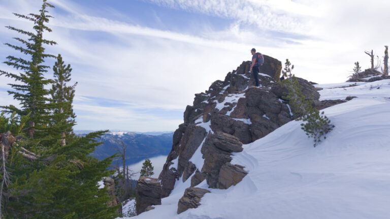 A hiker stands on a snowy rocky summit at Rifle Peak above Lake Tahoe