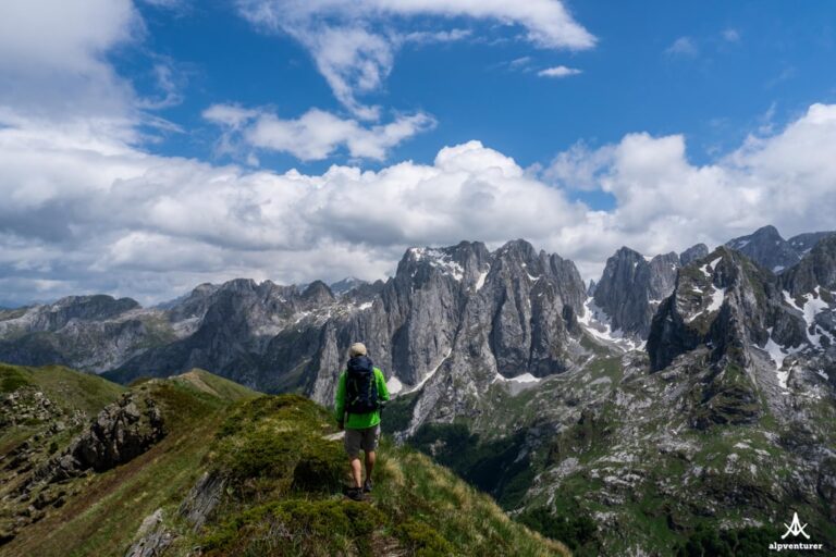A hiker on a grassy ridge above Lëpushë, facing rugged Albanian peaks beneath a cloudy blue sky