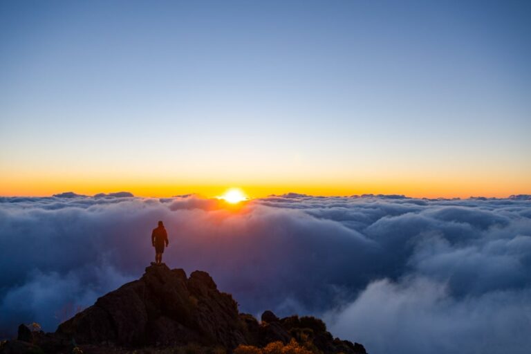 A hiker stands on a rocky summit above clouds as the sun rises over Madeira Island