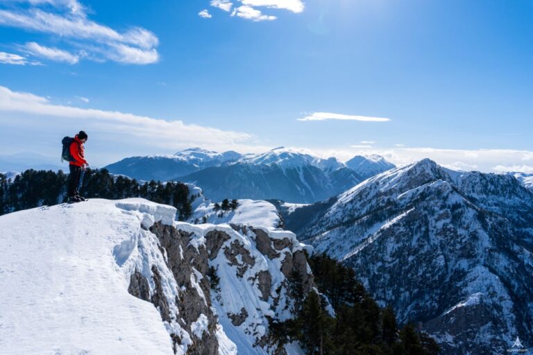 A hiker stands on a snowy ridge at Maja e Hasanit, overlooking forested Kosovo mountains
