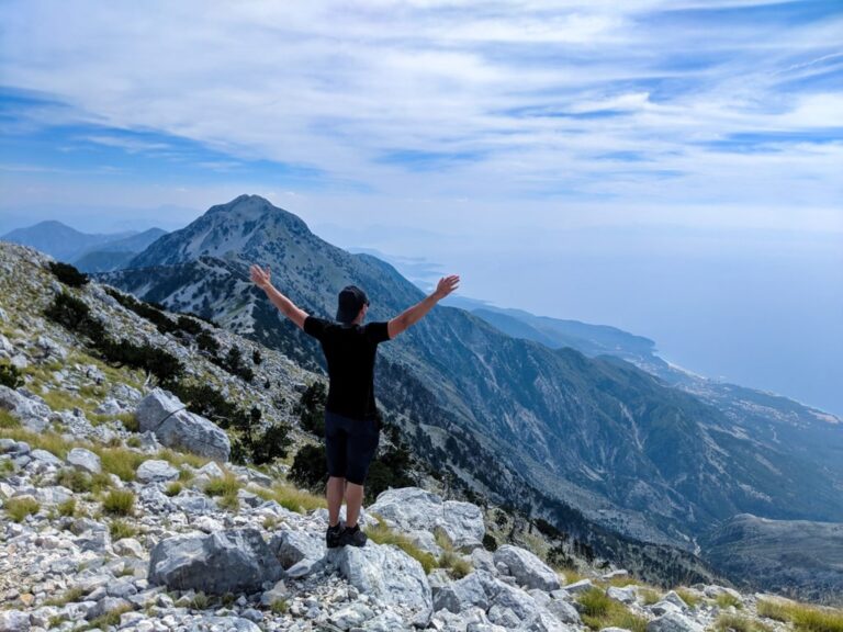 A hiker on the rocky summit of Maja e Qorres overlooks Albania's mountain ridges and distant coastline