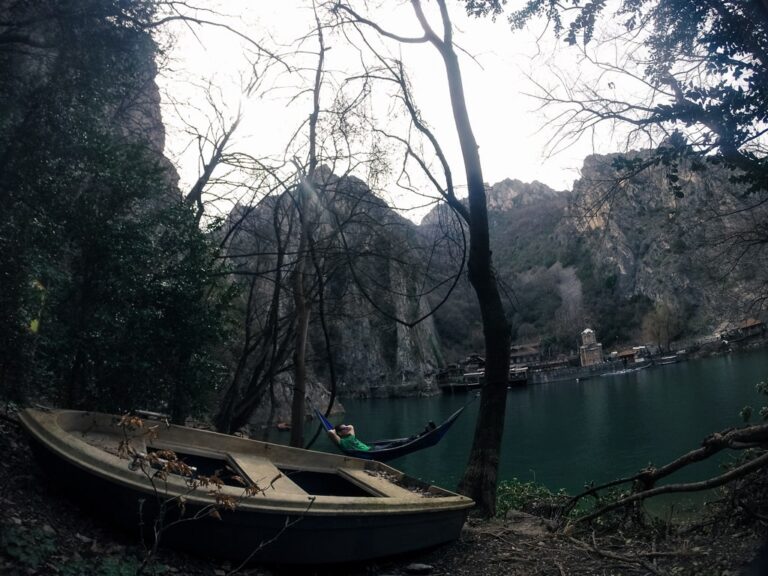 A small boat and hammock beside the green waters of Matka Canyon beneath bare trees and rocky cliffs