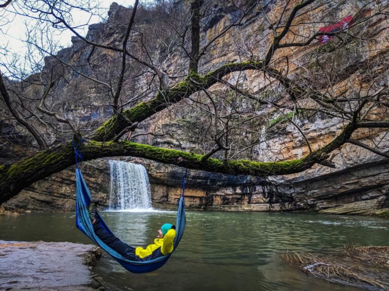 A hiker relaxes in a blue hammock beneath a waterfall at Mirusha, surrounded by bare trees and rocky cliffs