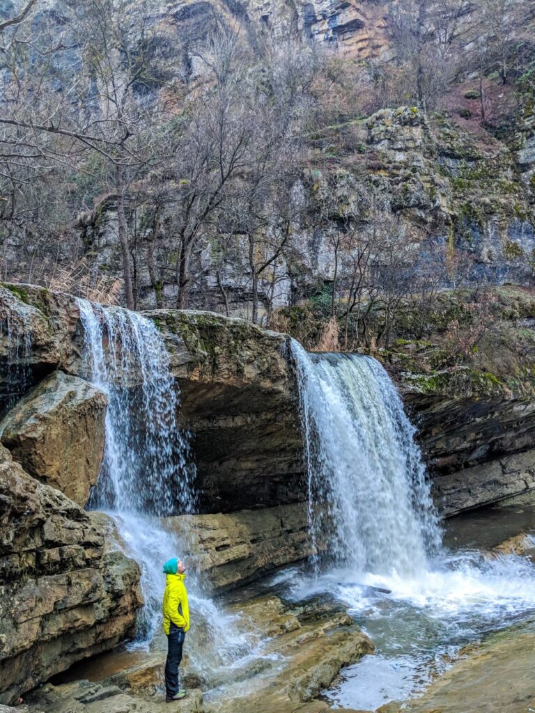 Two Mirusha waterfalls pour over layered rock beside a hiker in a bright yellow jacket
