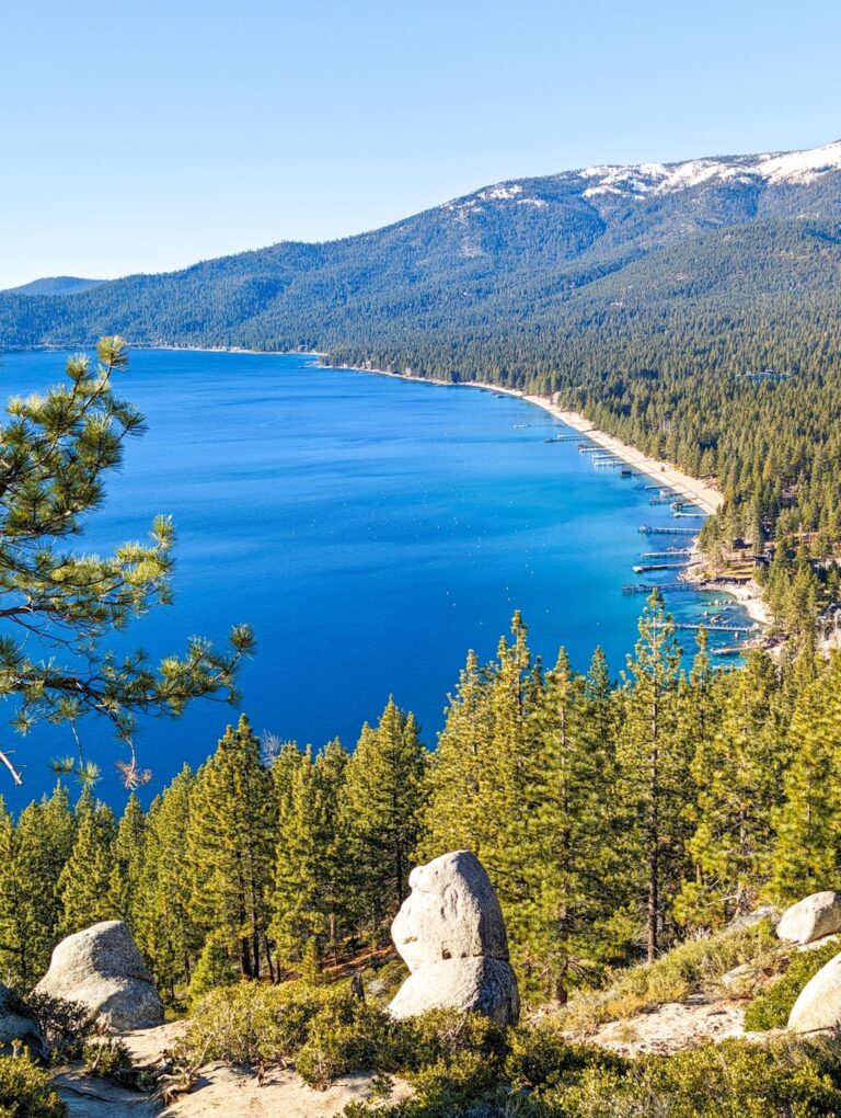 Monkey Rock overlooking the blue waters of Lake Tahoe, forested shoreline and snow-dusted mountains under clear skies