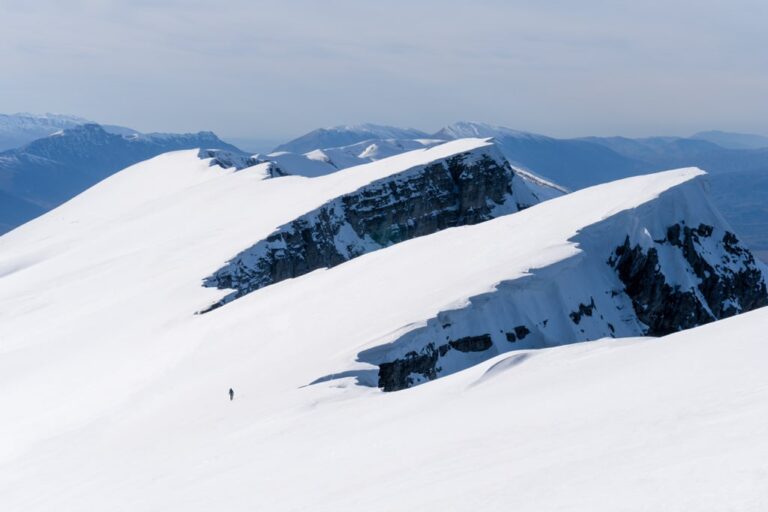A lone hiker crosses a broad snowy ridge on Mount Nemercke, with dark rocky cliffs and distant blue mountains
