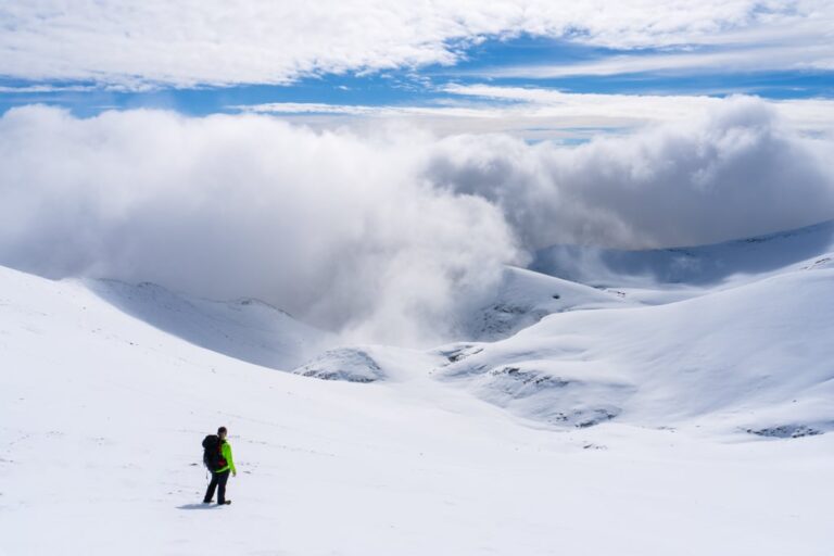 A hiker crosses a snowy slope on Mount Olympus as clouds roll over the surrounding peaks