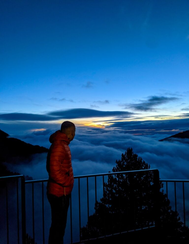 A hiker in an orange jacket looks over clouds from a railing on Mount Olympus at dusk
