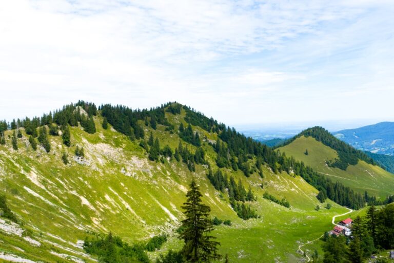 Green ridges and forested peaks along the Bodenschneid trail, with a small red-roofed building below
