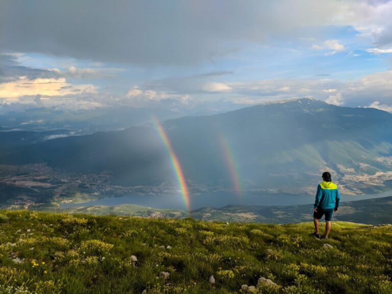 A hiker overlooks the Pashtrik mountain range beneath a double rainbow and cloudy evening sky