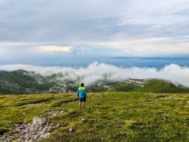 A hiker stands on a grassy, rocky slope above cloud-filled valleys on Pashtrik
