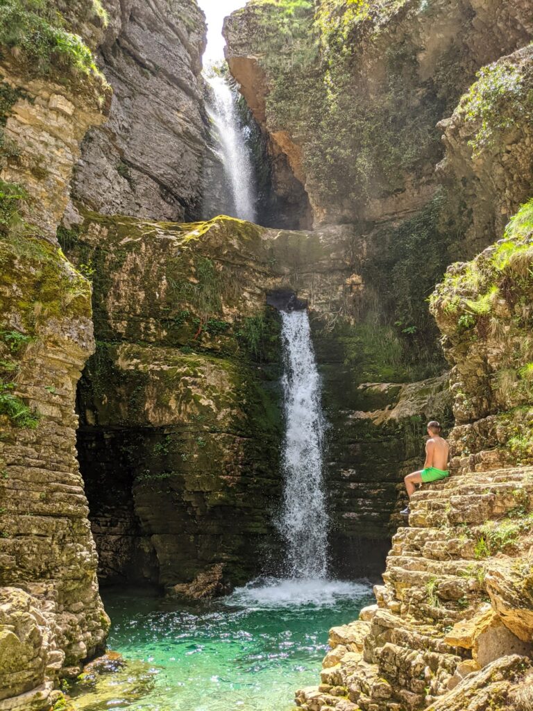 Peshtura Waterfall cascading into a turquoise pool between rocky canyon walls, with a hiker seated on a ledge