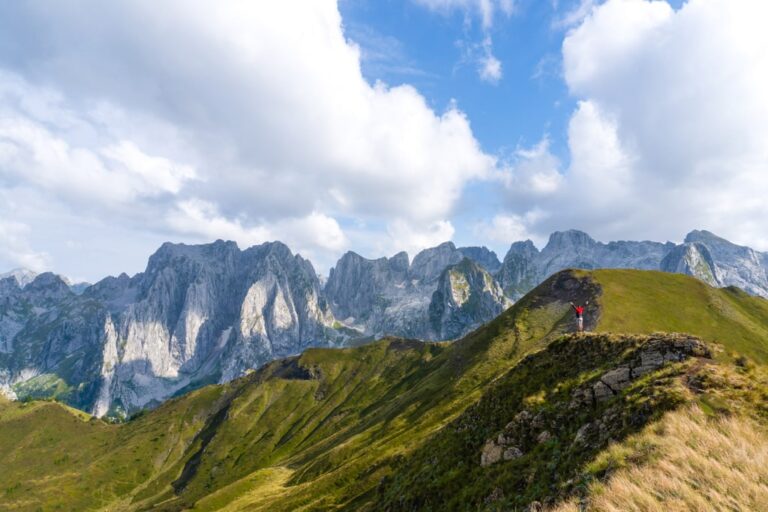 A hiker on a grassy ridge above Grebaje valley, with jagged Prokletije peaks beneath a cloudy blue sky