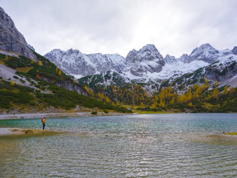 A hiker stands by Seebensee, with autumn trees and snow covered peaks under a cloudy sky