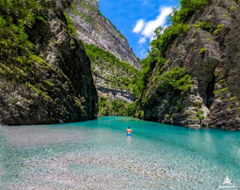 A swimmer in the turquoise Shala River beneath steep green cliffs on a sunny day