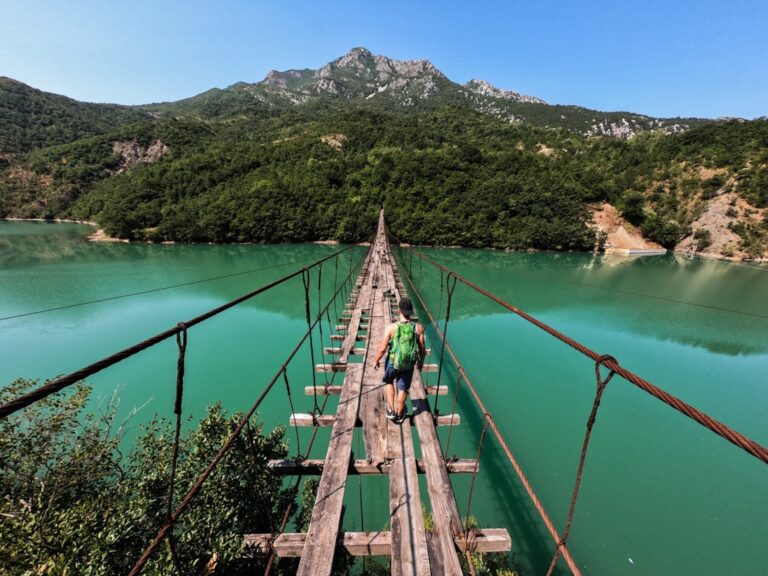 A hiker crosses a narrow suspension bridge over turquoise Shkopet Lake, with forested hills and mountains beyond