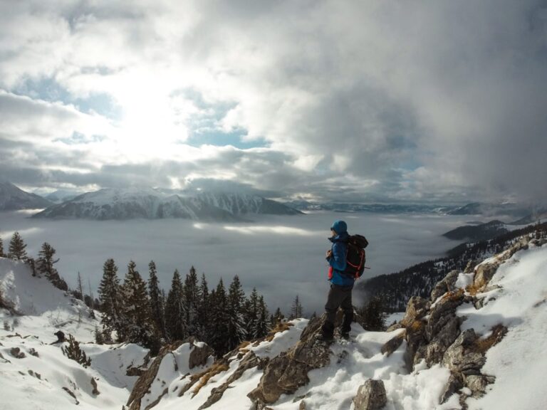 A hiker overlooks the snow-covered Rugova Mountains from a rocky, pine-lined slope in winter