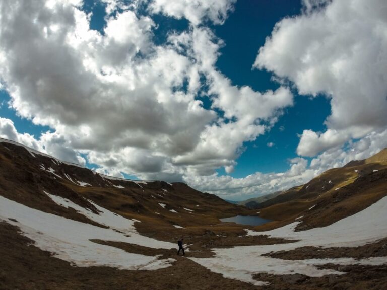 A hiker crosses a snow-patched valley toward Shutman Lake beneath a cloudy sky in the Sharr Mountains
