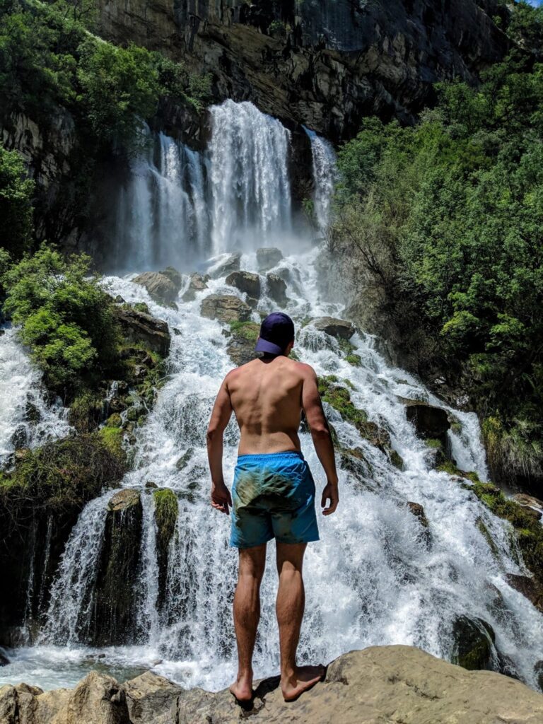 A hiker stands barefoot before Sotira Waterfalls, where spring water cascades over rocks beneath a forested cliff