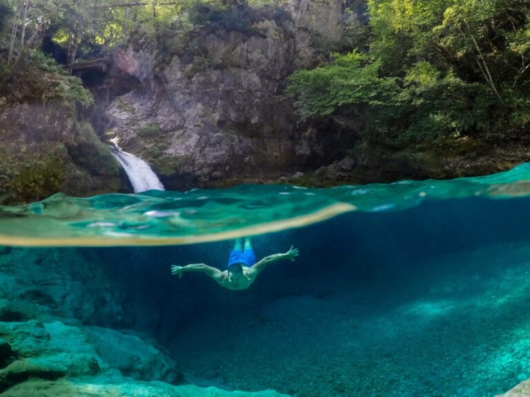 A swimmer beneath turquoise water below a small waterfall in Theth National Park