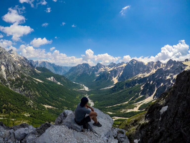 A hiker rests on rocks overlooking Valbona Valley and its rugged peaks under a bright blue summer sky