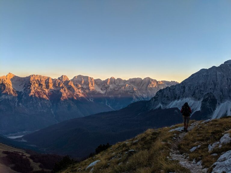 A hiker on a rocky trail above the Valbona Valley, with mountain peaks lit by early morning light